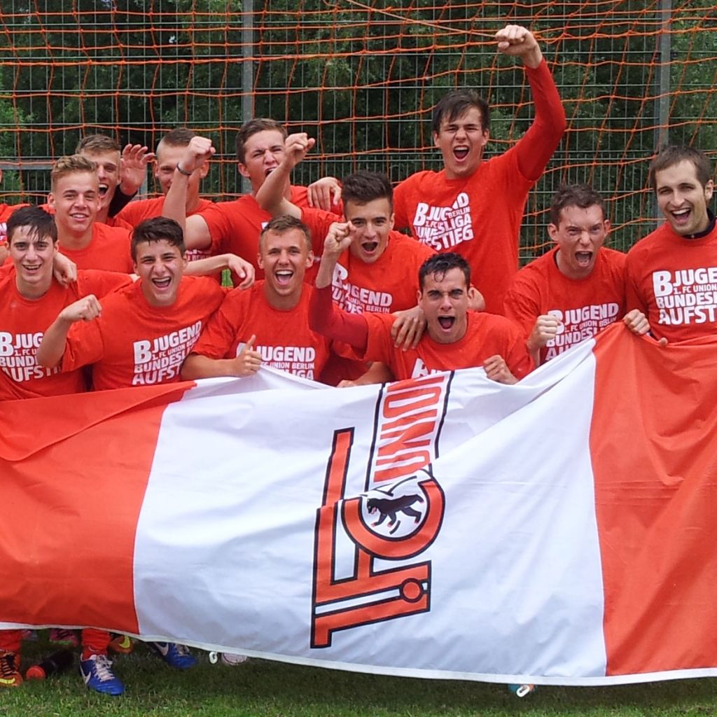 Eine Gruppe junger Fußballspieler in roten T-Shirts posiert freudig vor einem großen Banner mit ihrem Teamlogo.