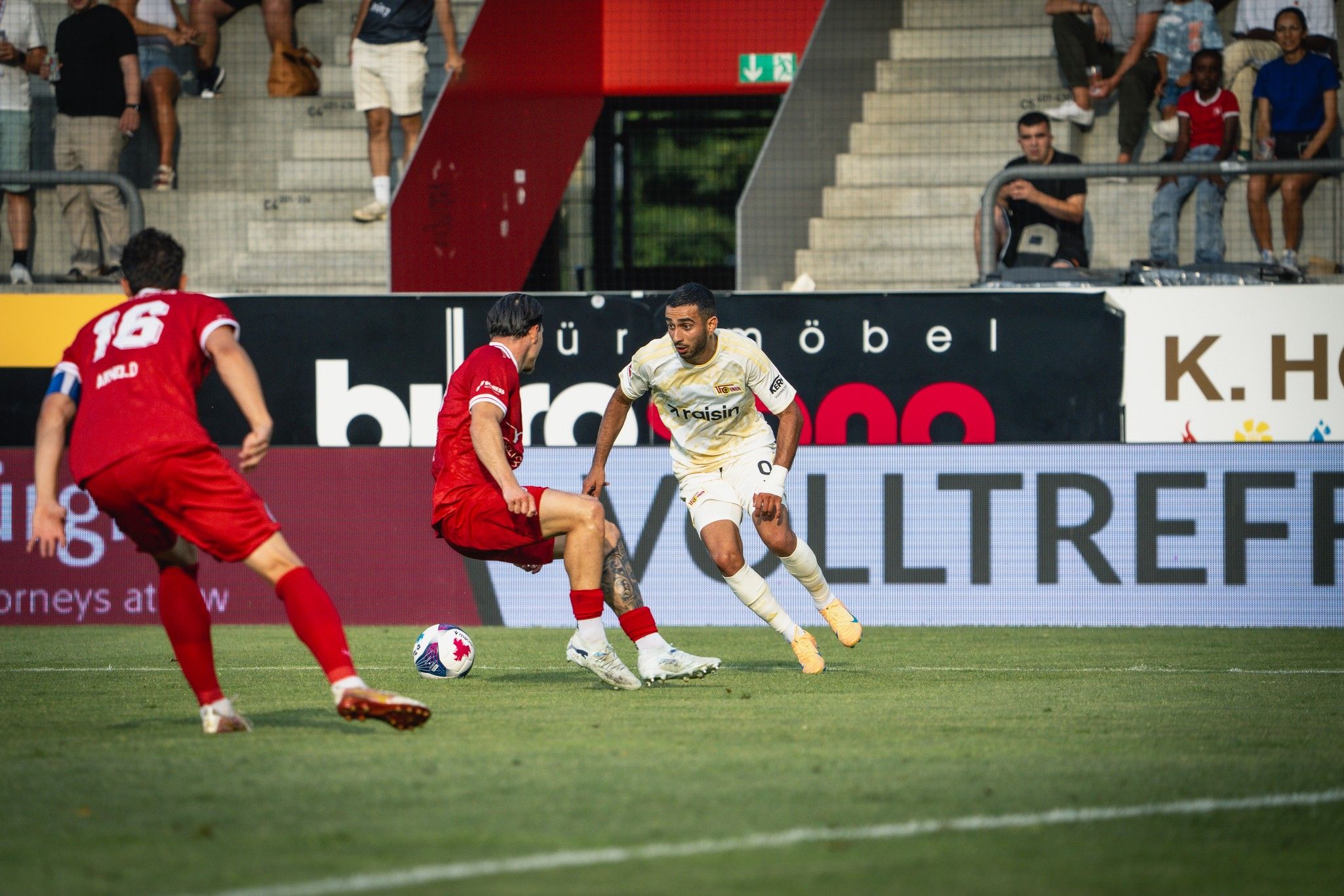 Fußballspiel mit Spielern in roten und weißen Trikots auf dem Spielfeld, Zuschauer im Hintergrund.