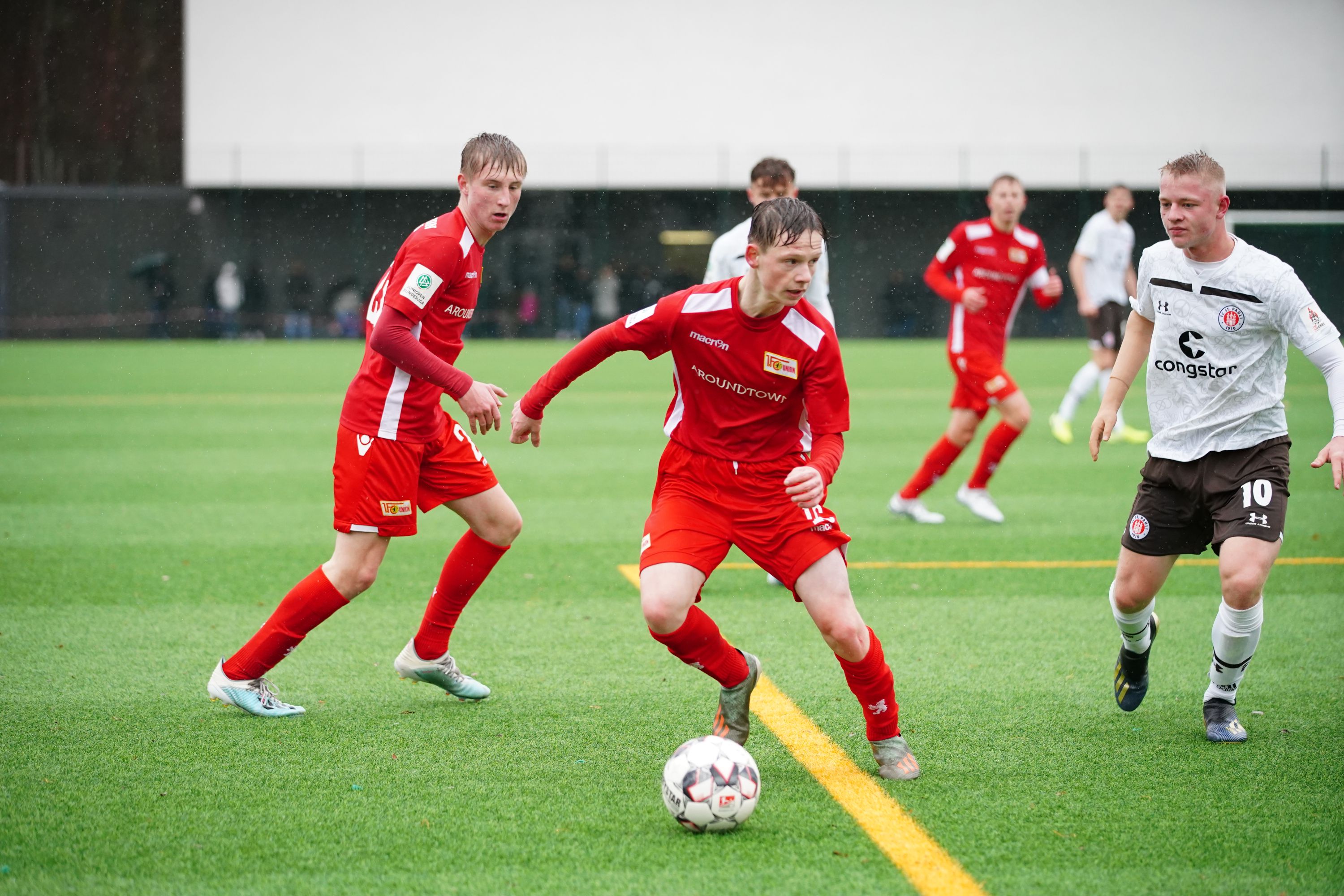 Zwei Spieler in roten Trikots dribbeln den Ball über ein grünes Fußballfeld, während ein gegnerischer Spieler in Weiß näherkommt.