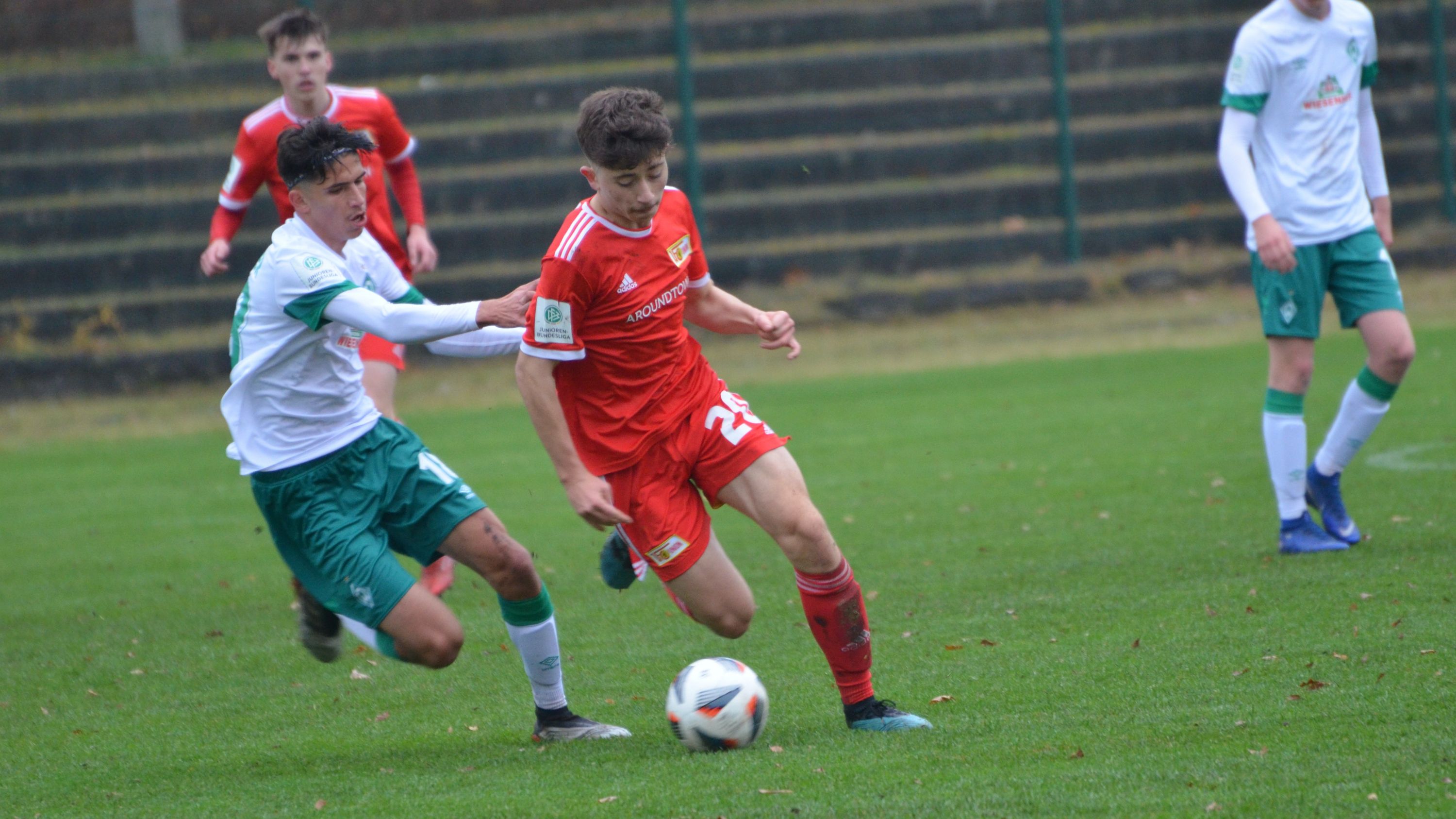 Ein Fußballspiel, bei dem ein Spieler in roten Trikots den Ball führt, während ein Spieler in weißen und grünen Trikots ihn verfolgt.