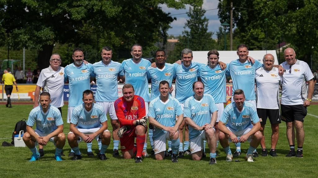 Eine Fußballmannschaft in hellblauem Trikot steht auf einem Rasenfeld, umgeben von Bäumen und mit einem klaren Himmel im Hintergrund.