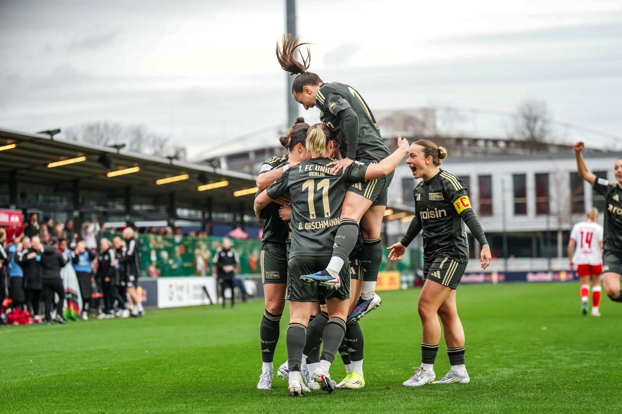 Fußballspielerinnen feiern einen Torerfolg auf dem Spielfeld, umgeben von jubelnden Teamkollegen und Zuschauern im Hintergrund.