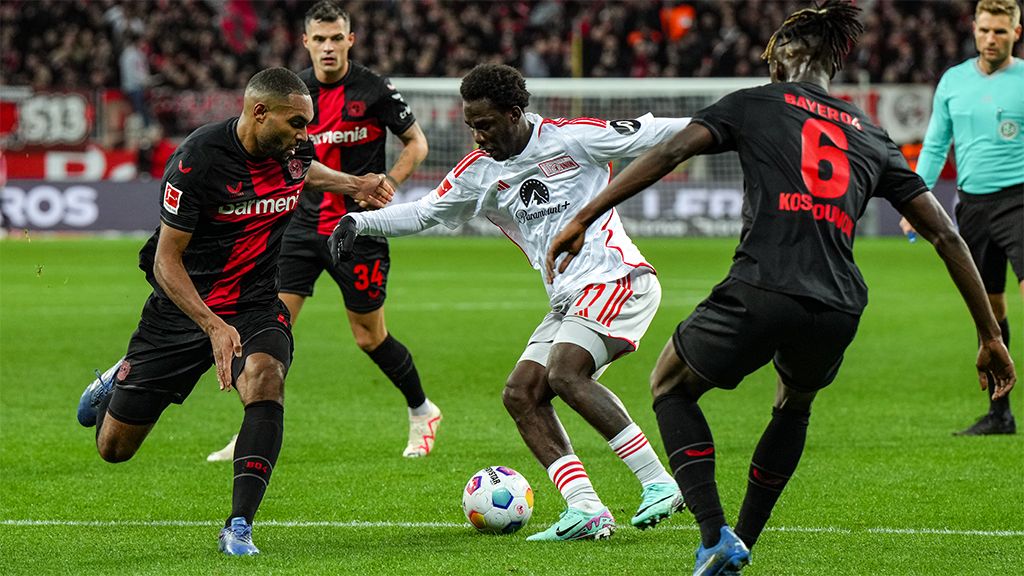 Ein Fußballspieler im weißen Trikot kämpft um den Ball gegen zwei Spieler im schwarzen Trikot auf einem Stadionfeld.
