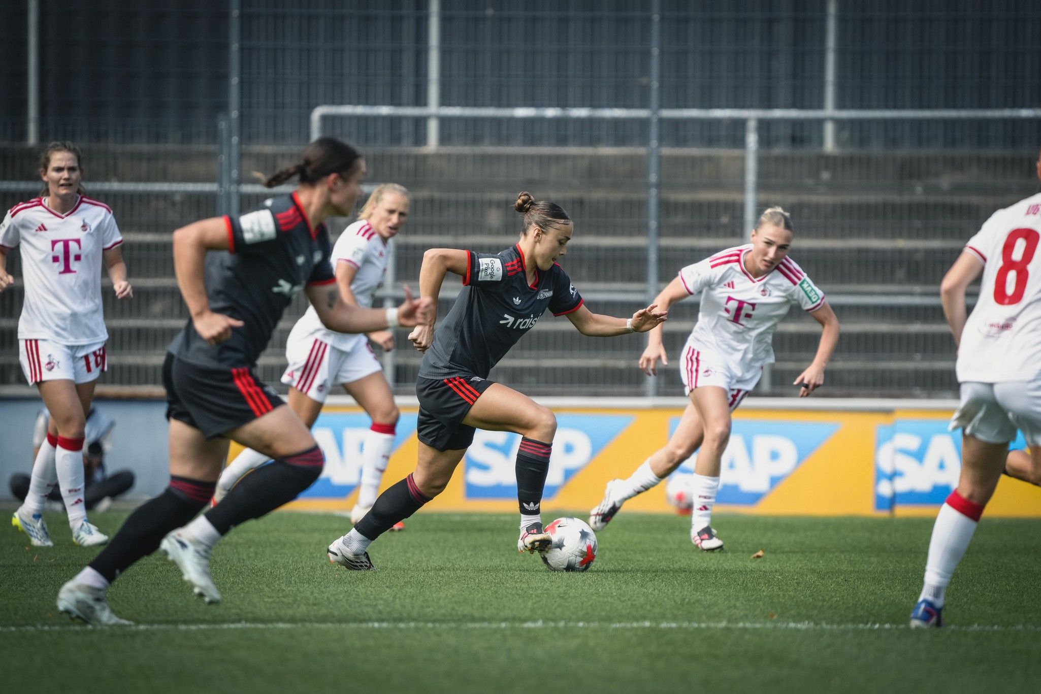 Eine Fußballspielerin in schwarzer Uniform dribbelt den Ball, während Gegnerinnen in weißen Trikots versuchen, sie zu stoppen.