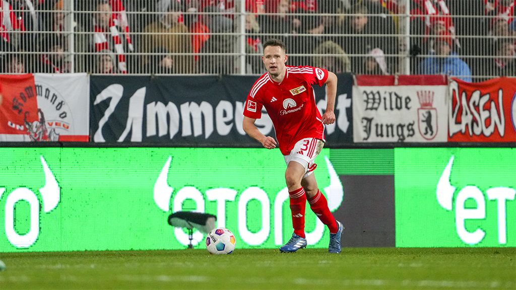 Ein Fußballspieler in roter Uniform sprintet über das Spielfeld, während Fans im Hintergrund ein Banner halten.