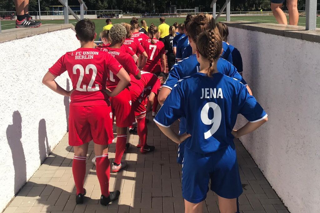 Zwei Fußballmannschaften in roten und blauen Trikots betreten das Spielfeld durch einen Tunnel.