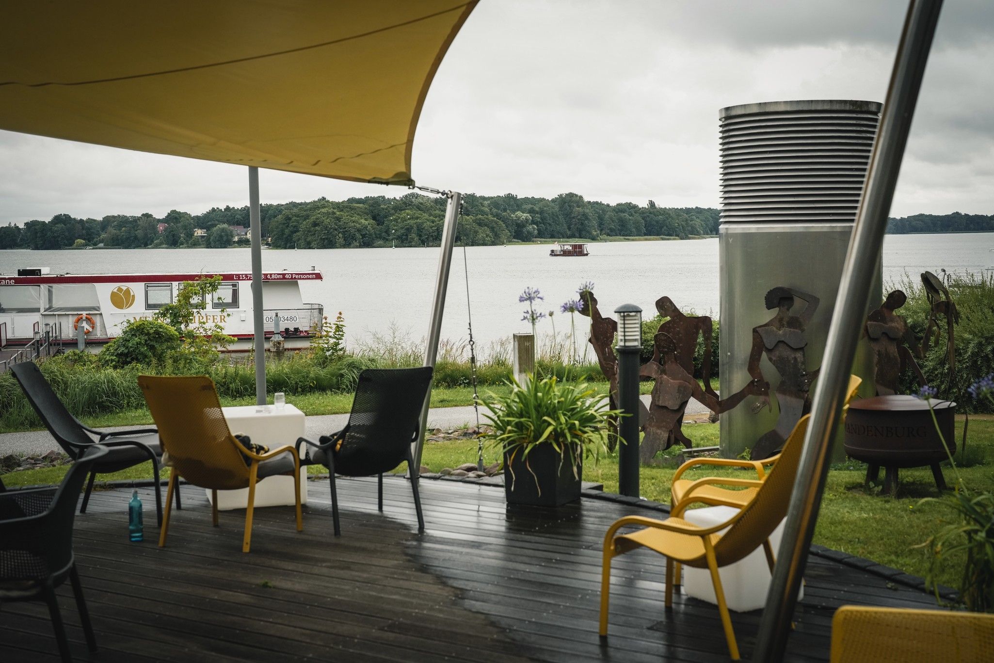 Überdachte Terrasse mit Stühlen, Blick auf einen See und einen vorbeifahrenden Bootsfahrzeug, umgeben von Pflanzen und Kunstskulpturen.