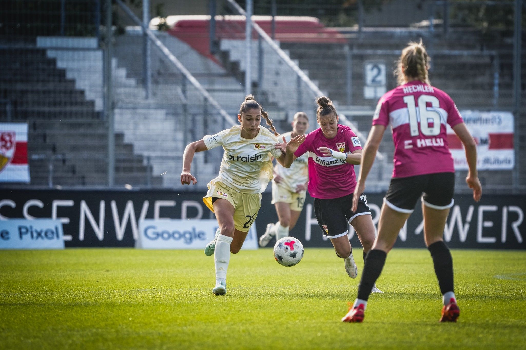 Eine Spielerin in gelb läuft mit dem Ball, während eine Torhüterin in pink sie verfolgt. Im Hintergrund sind Zuschauer und andere Spielerinnen.