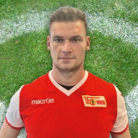 Ein Fußballspieler mit kurzem, blondem Haar trägt ein rotes Trikot mit dem Logo des 1. FC Union auf einem grasbewachsenen Feld.