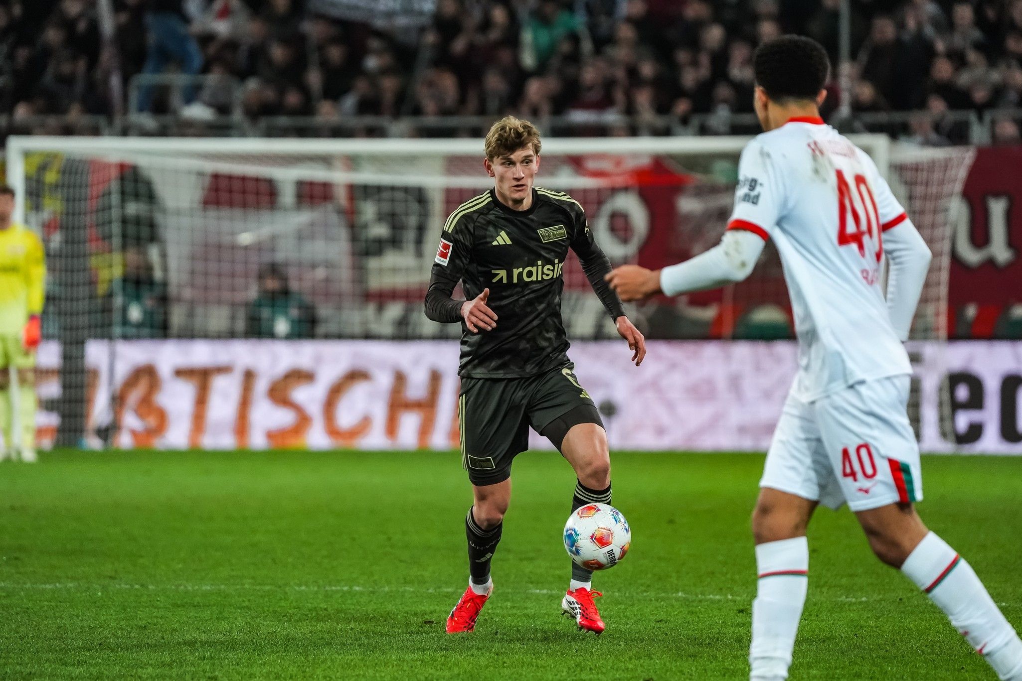 Ein Fußballspieler in schwarzer Ausrüstung dribbelt den Ball, während ein anderer Spieler in weißer Ausrüstung ihn beobachtet.