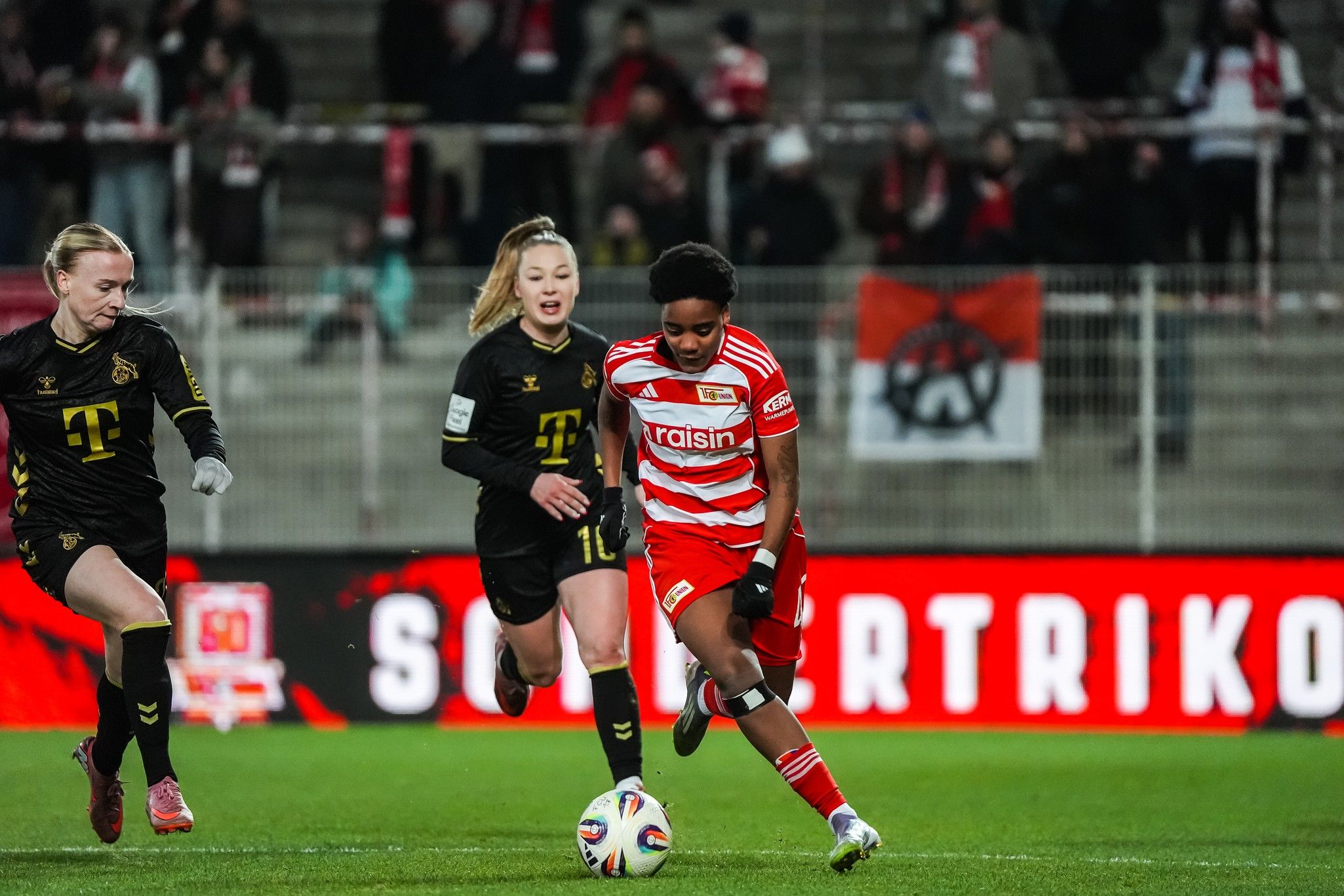 Eine Fußballspielerin in rot-weißer Uniform dribbelt den Ball, während zwei Gegnerinnen in schwarz-gelben Trikots verfolgen.