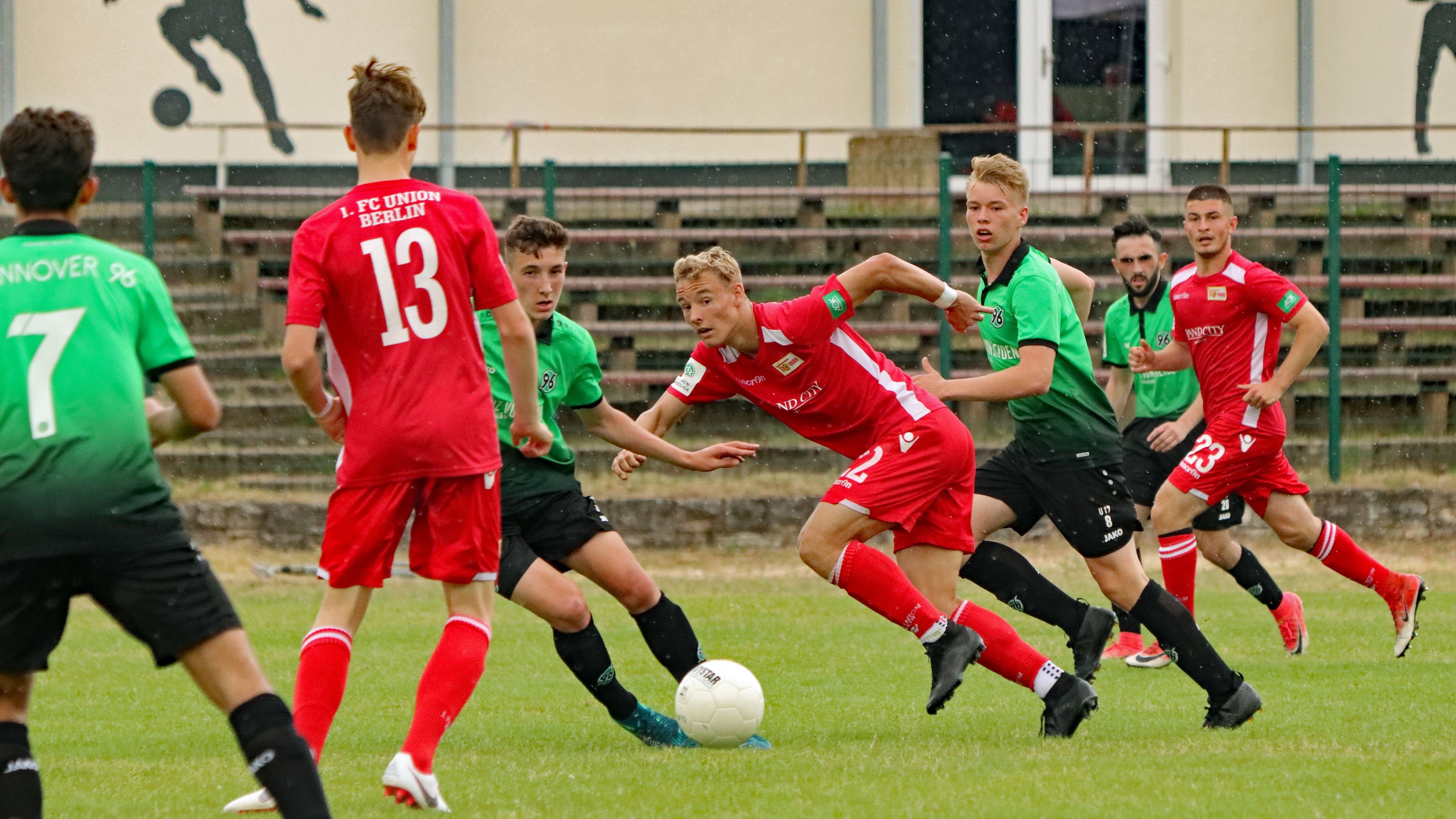 Ein Fußballspiel für Jugendliche. Die Spieler haben rote und grüne Trikots. Sie kämpfen um den Ball. Im Hintergrund sind Zuschauer.