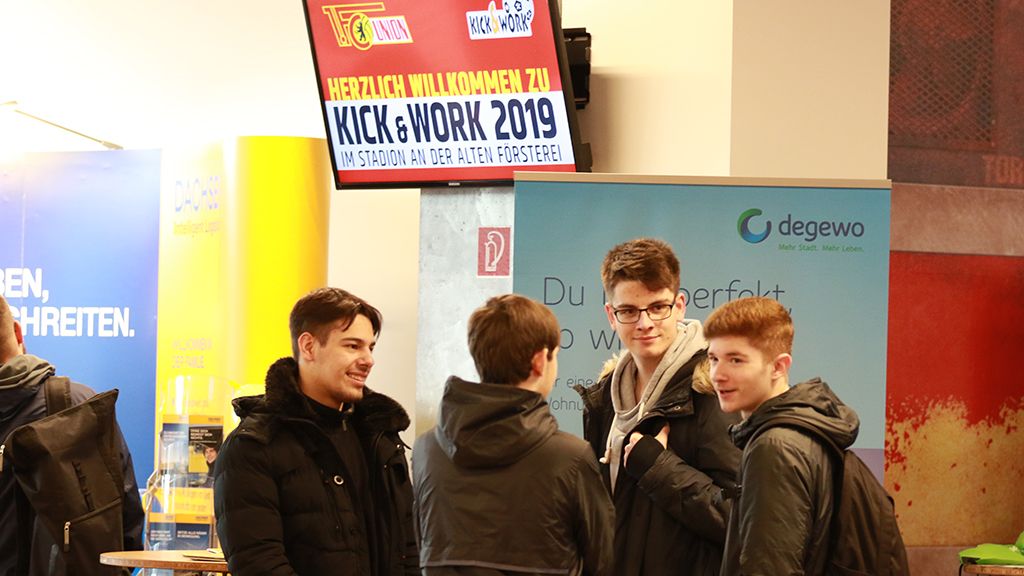 Gruppengespräch junger Menschen in einem Foyer mit Informationsständen und Werbeplakaten.