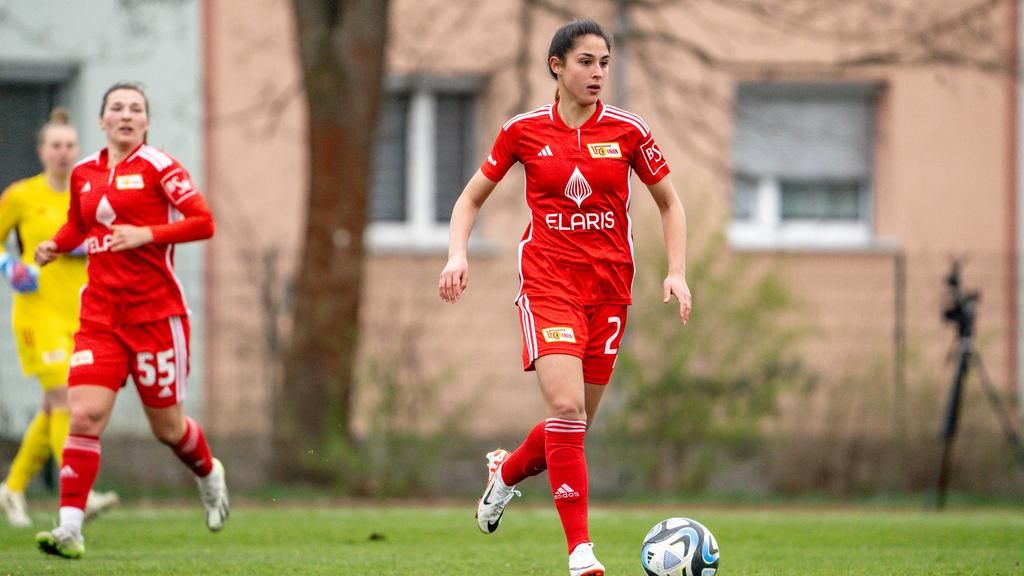 A female soccer player in a red jersey dribbles the ball while two teammates run in the background.