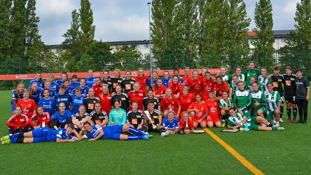 Gruppenshot von Fußballspielerinnen in verschiedenen Trikots auf einem Rasenplatz, umgeben von Bäumen und einem klaren Himmel.