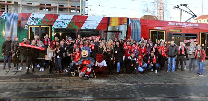 Gruppierung von Menschen in Fankleidung vor einer Straßenbahn, einige halten Banner und Schilder hoch.