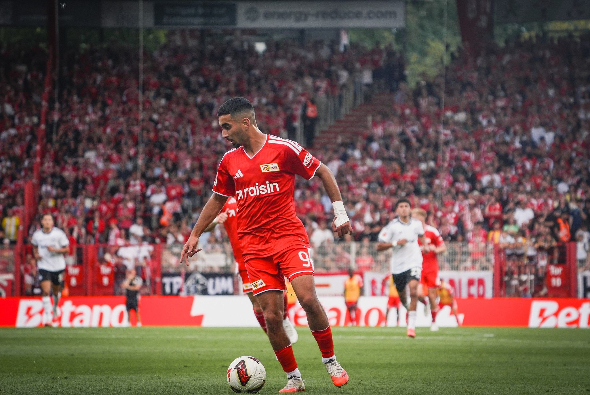 Ein Fußballspieler in roter Trikotfarbe dribbelt den Ball auf einem sportlichen Platz, umgeben von jubelnden Fans.