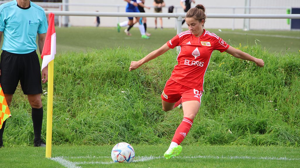 A player in a red jersey takes a corner kick while a referee in blue watches.