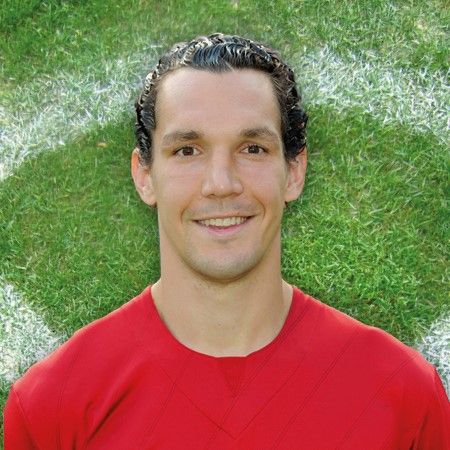 Portrait eines lächelnden Fußballspielers in rotem Trikot, vor einem grünen Rasenhintergrund.
