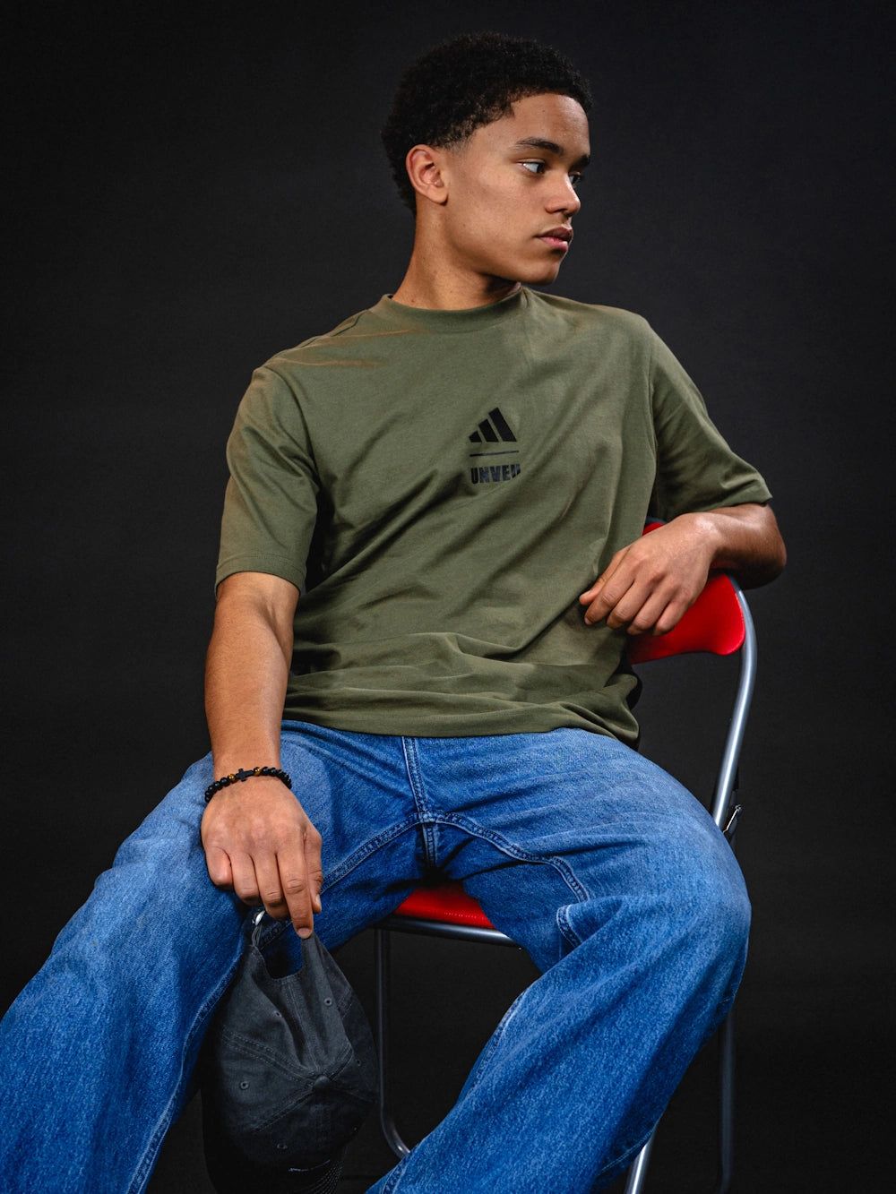 Junger Mann sitzt auf einem roten Stuhl, trägt ein grünes T-Shirt mit Adidas-Logo und Jeans, schaut nachdenklich zur Seite.