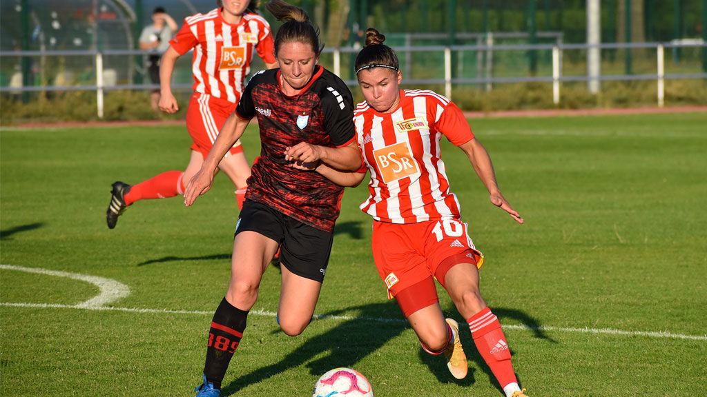 Zwei Frauenfußballerinnen kämpfen um den Ball auf einem Sportplatz. Beide tragen Trikots in unterschiedlichen Farben.