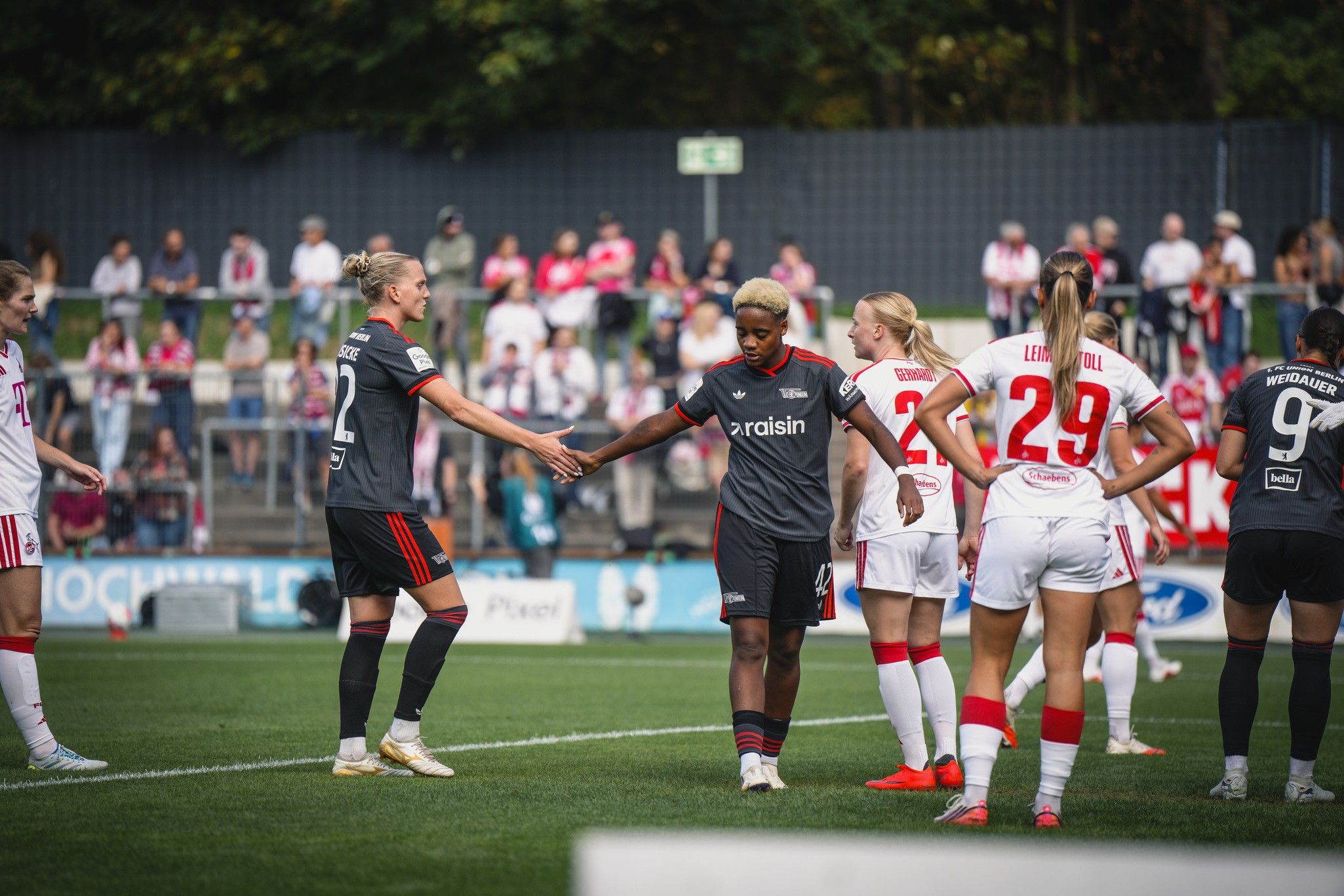 Zwei Fußballspielerinnen stehen auf dem Platz und schütteln sich die Hände, während andere Spielerinnen im Hintergrund stehen.