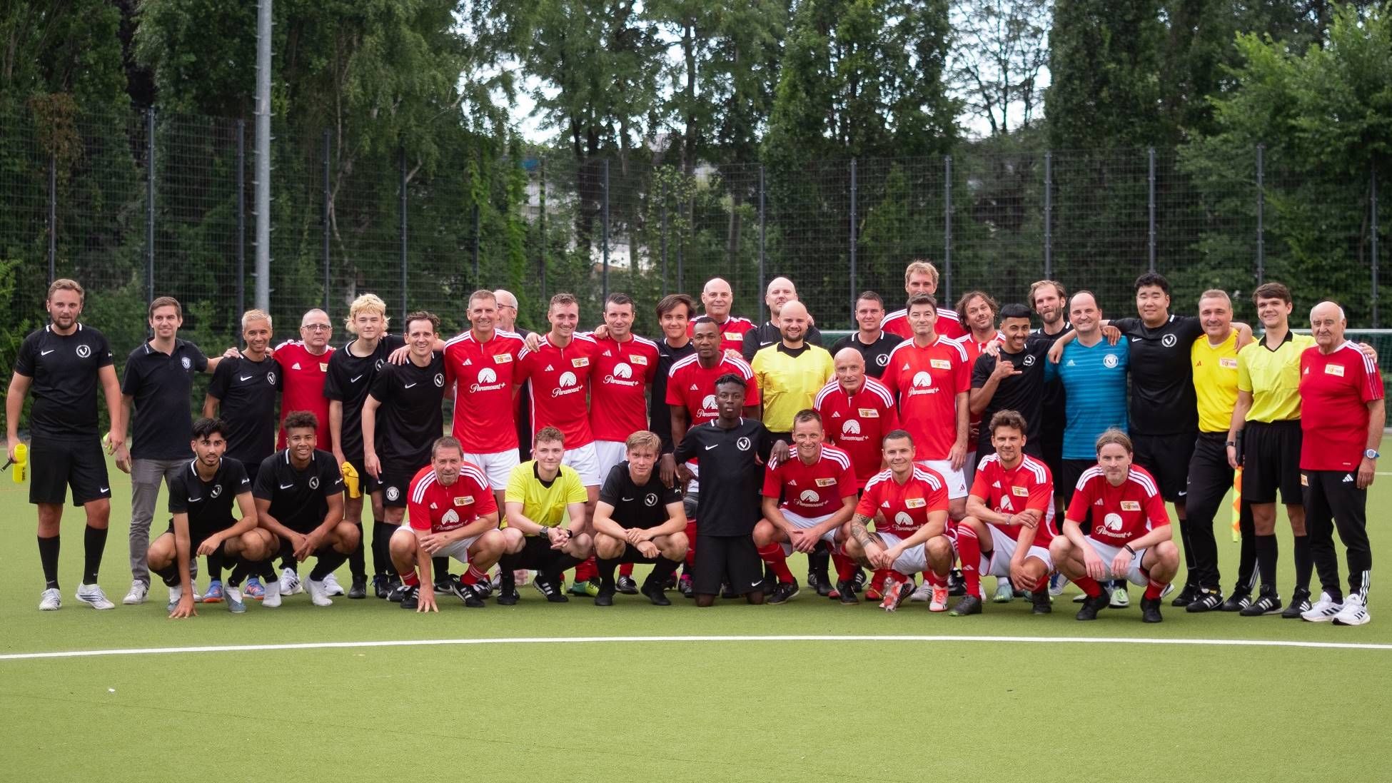 Gruppenfoto von mehreren Menschen in roten und schwarzen Fußballtrikots, die lächeln und auf einem Sportplatz stehen.