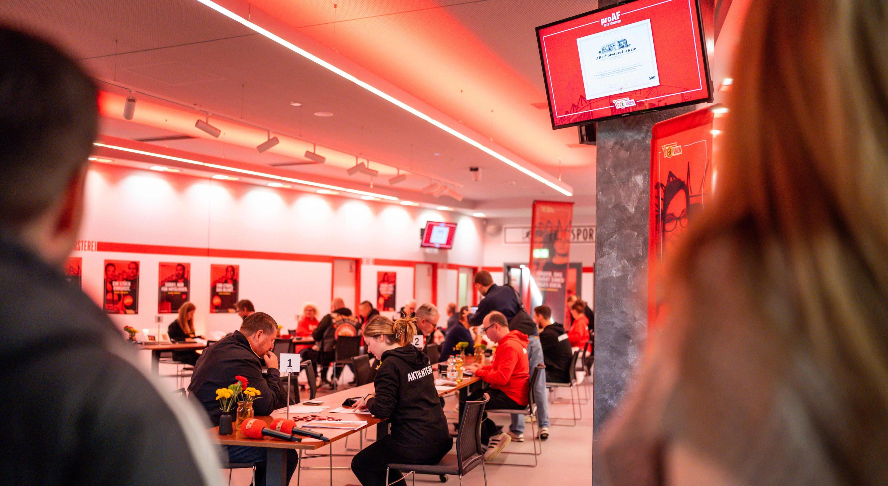 Menschen sitzen an Tischen in einem modernen Raum, während ein Bildschirm Informationen anzeigt. Rote Beleuchtung sorgt für Atmosphäre.