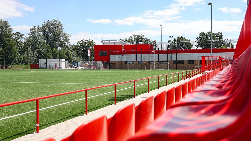 Empty red stadium benches are at the side of a well-maintained football field under a blue sky.