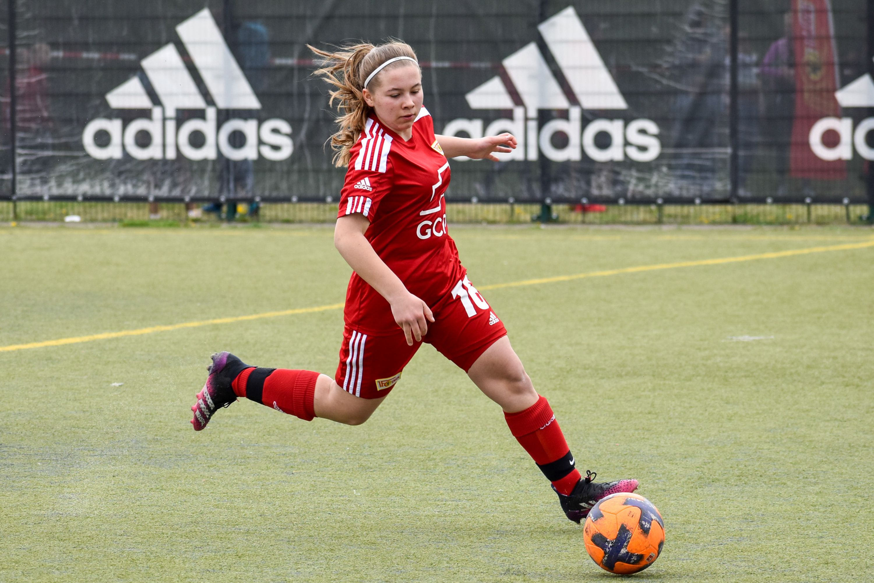 Ein Mädchen in rotem Fußballtrikot tritt einen Ball auf einem Kunstrasenplatz. Hintergrund mit Adidas-Werbung.