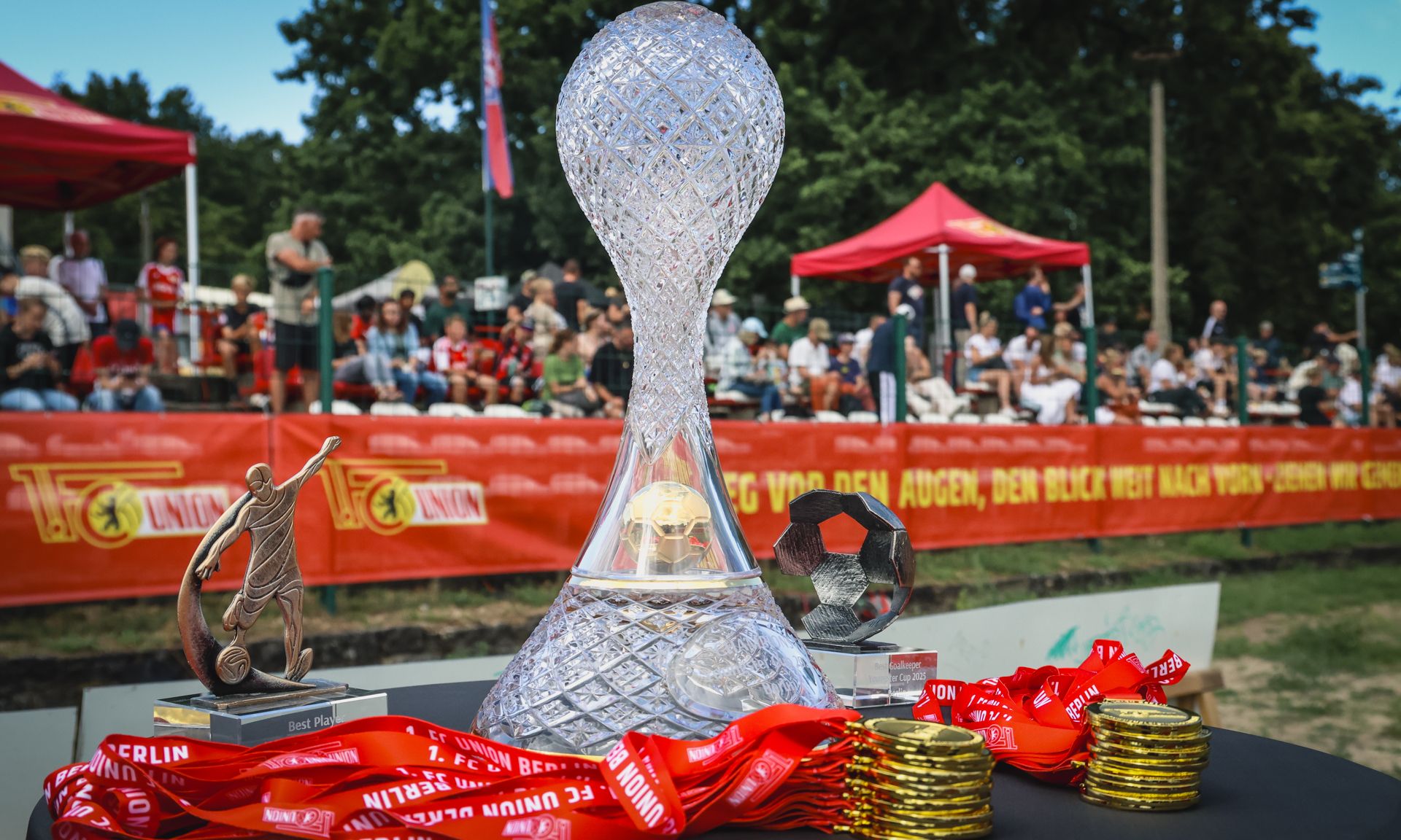 Ein gläserner Pokal steht auf einem Tisch, umgeben von Medaillen und einer Trophäe, mit Zuschauern im Hintergrund.