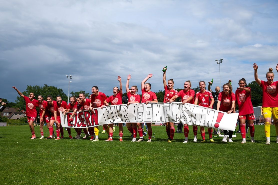 Fußballmannschaft in roten Trikots jubelt auf dem Platz, hält ein Banner mit der Aufschrift „LEISTERN NUR GEMEINSAM!“ hoch.