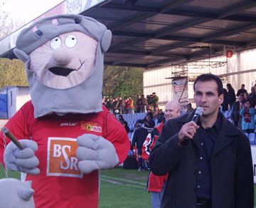 Maskottchen mit grauem Bart in rotem Trikot steht neben einem Mann mit Mikrofon auf einem Sportfeld. Zuschauer im Hintergrund.