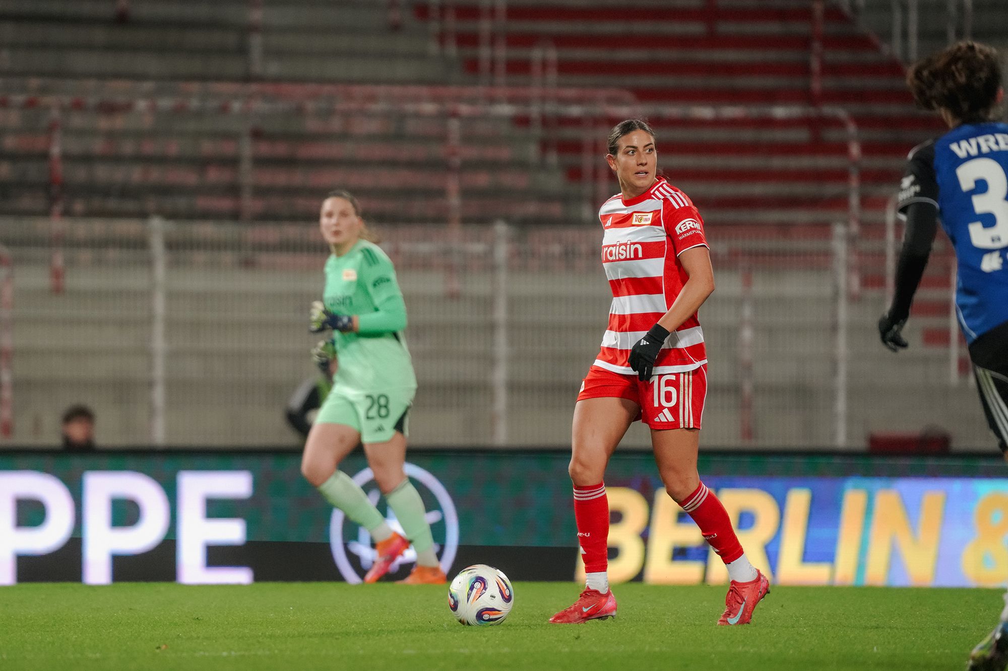 Fußballspiel, Spielerin mit der Nummer 16 in rotem Trikot, Torhüterin im Hintergrund, Stadionbeleuchtung, Abendstimmung.