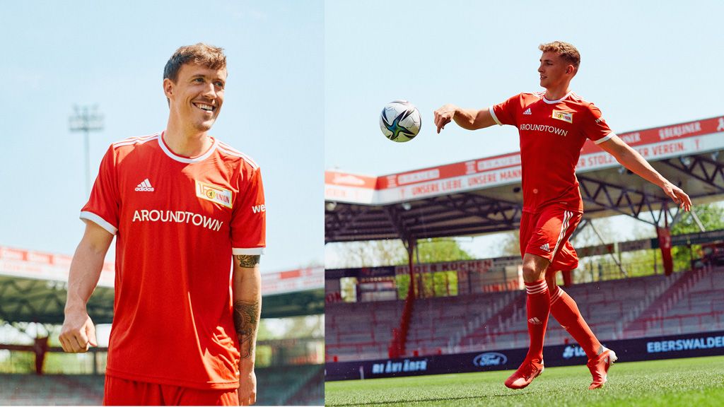 Zwei Fußballspieler in roten Trikots stehen im Stadion, einer lächelt, der andere jongliert den Ball.