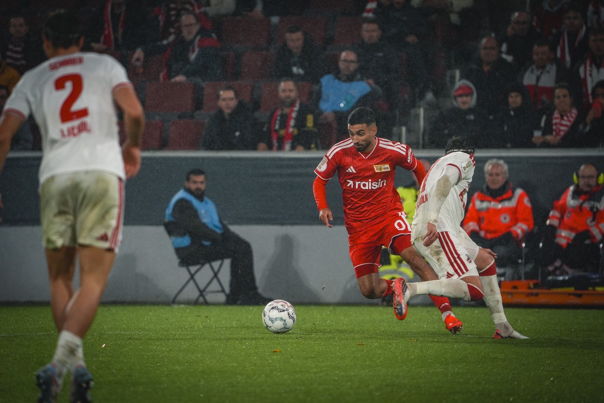 Spielszene mit einem Fußballspieler in rotem Trikot, der im Wettlauf um den Ball von einem gegnerischen Spieler verfolgt wird.