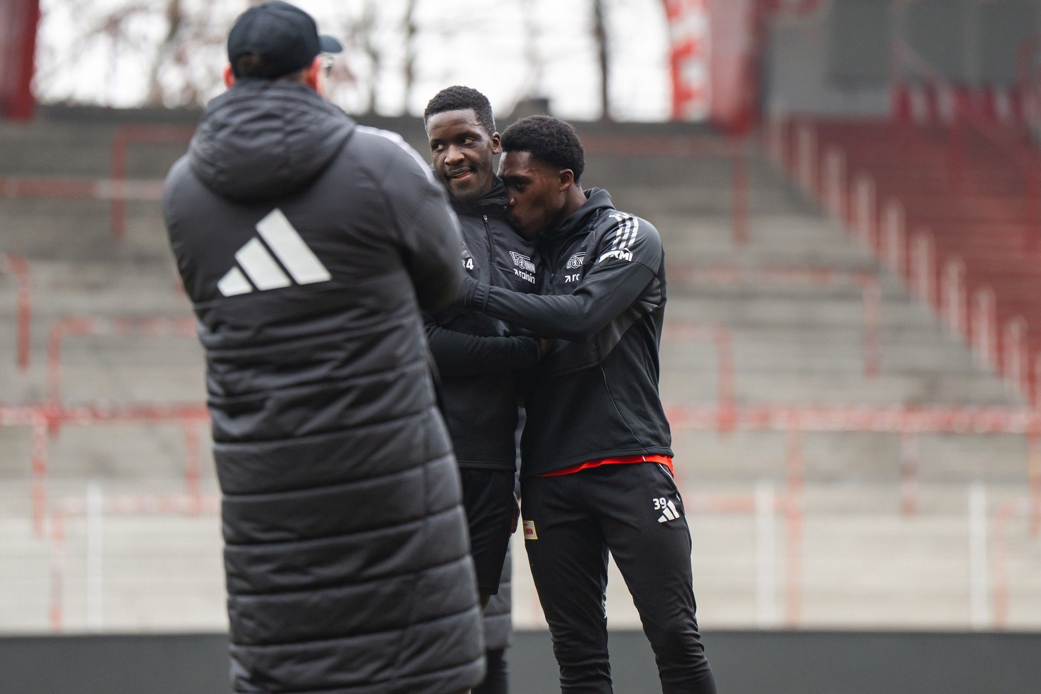 Drei Personen in Trainingsanzügen stehen in einem Stadion, zwei umarmen sich freundlich, während eine Person ihnen gegenüber steht.