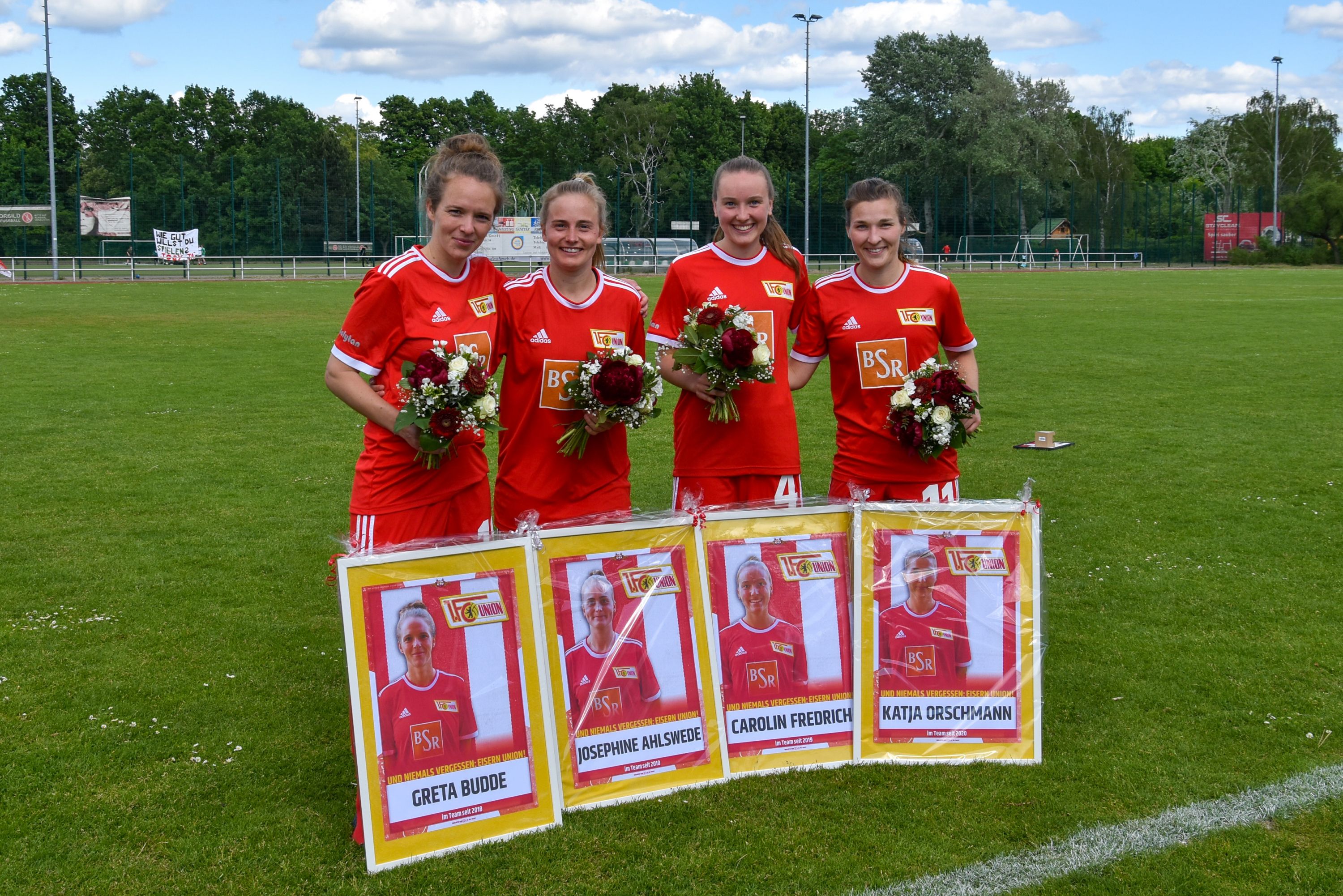 Vier Fußballspielerinnen in roten Trikots stehen auf einem Rasenplatz, halten Blumensträuße und posieren vor ihren Plakaten.