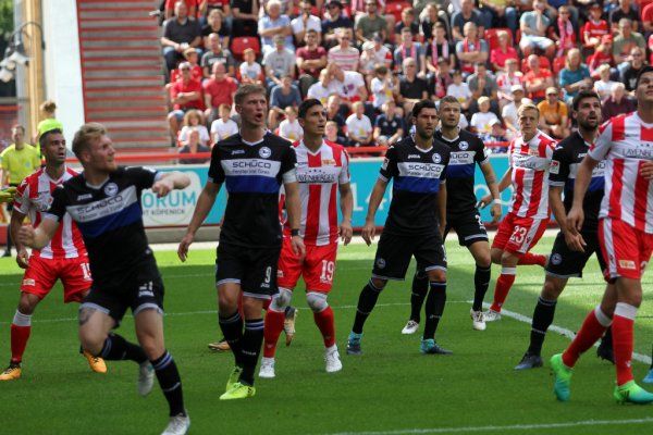 Football players in black-and-white and red-and-white jerseys during a game on the field, spectators in the background.