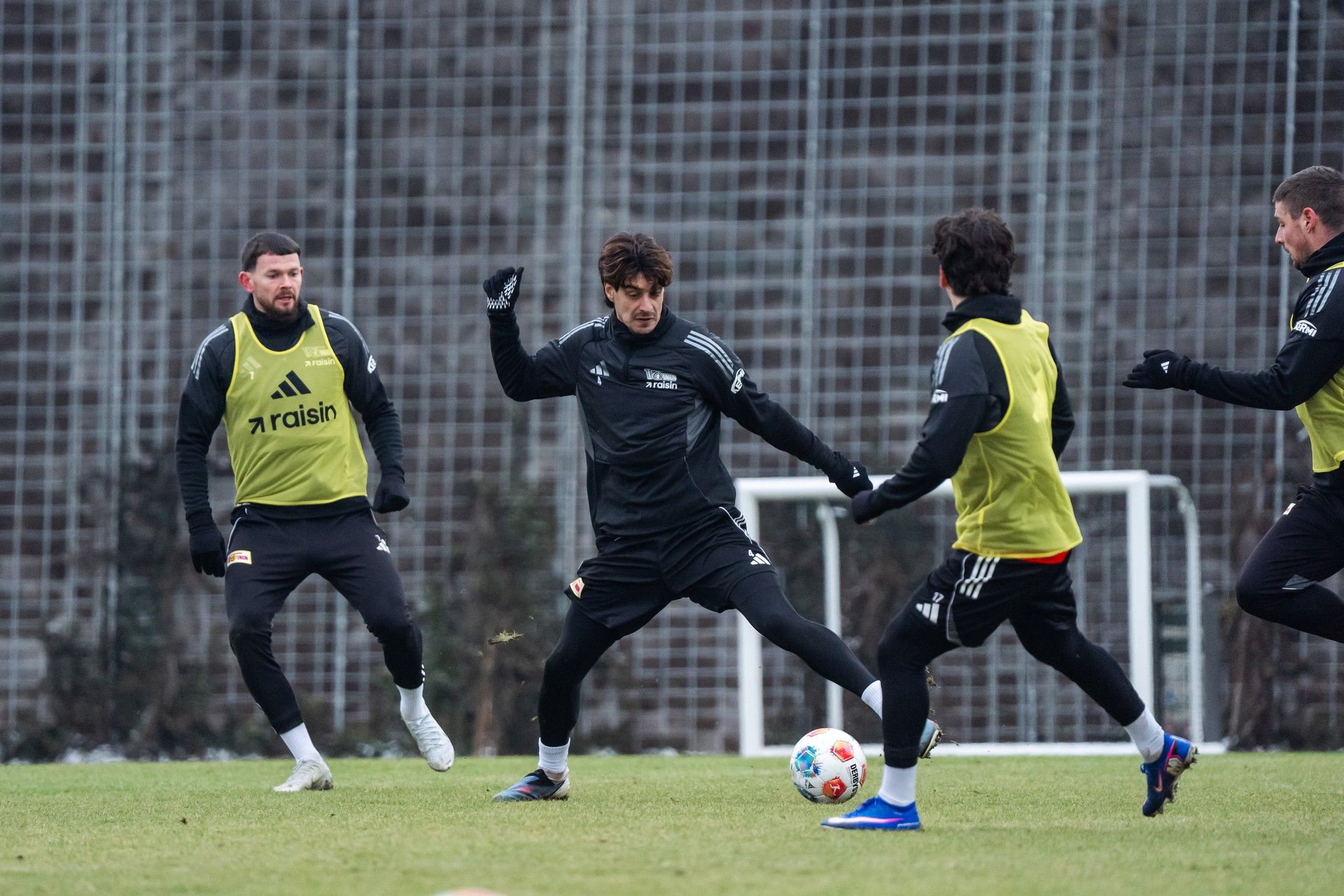 Fußballspieler in Trainingskleidung üben auf dem Platz, während einer den Ball dribbelt. Hintergrund mit Tor und Zaun.