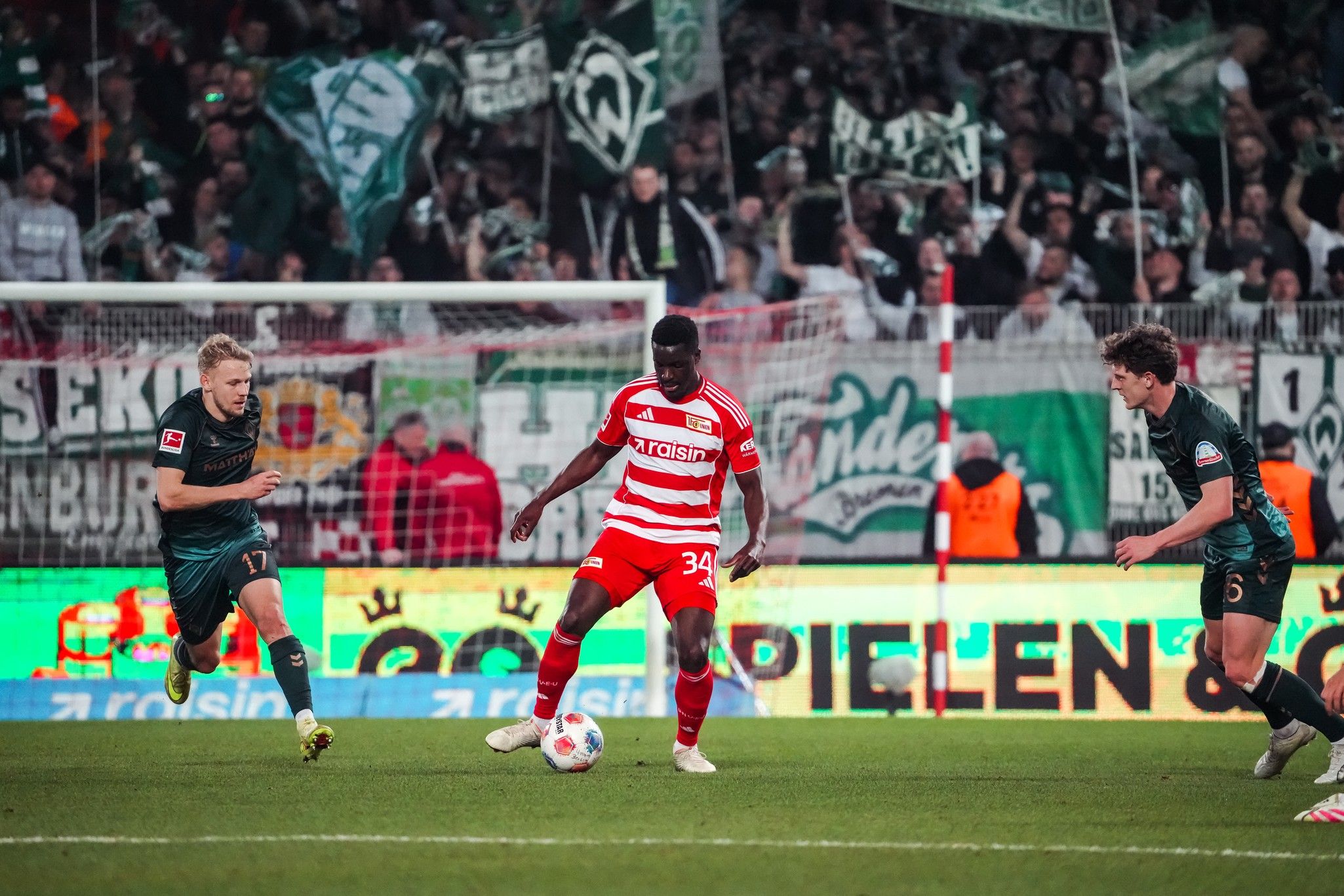 Spielszene aus einem Fußballspiel mit zwei Spielern in dunkler und roter Uniform, die den Ball im Stadion verfolgen.