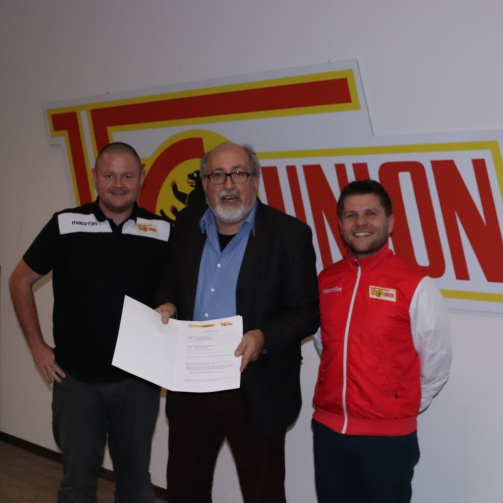 Drei Männer halten ein Dokument, lächeln und stehen vor einem großen Union-Logo an einer Wand.