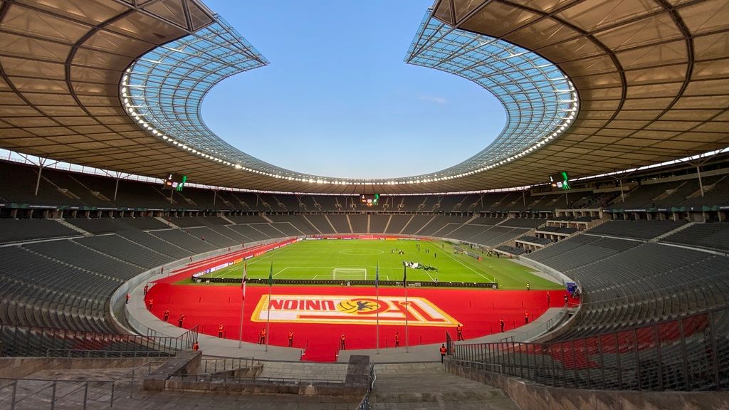 Innenansicht eines Stadion mit grünem Spielfeld, roten Akzenten und einer modernen Dachkonstruktion, bei klarem Wetter.