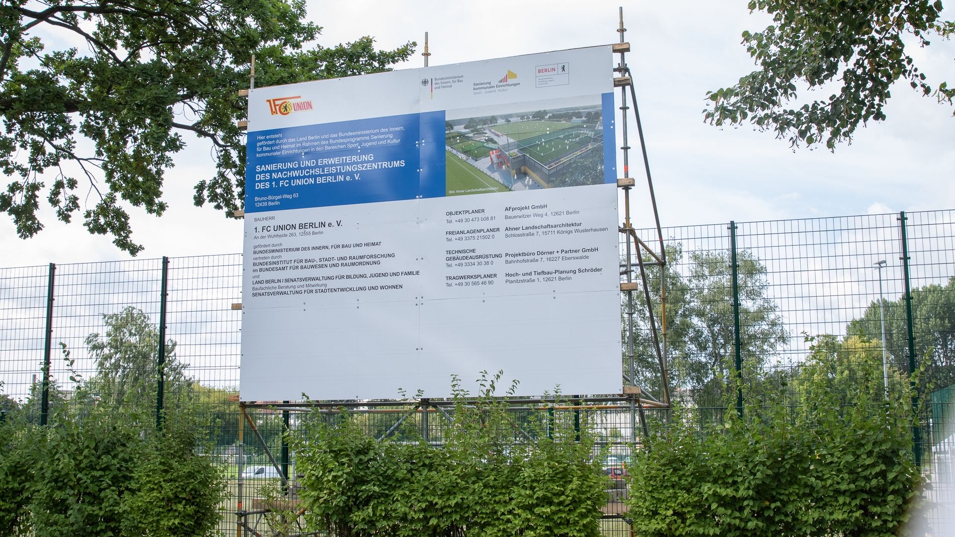 Baustellenschild mit Informationen zu einem Neubauvorhaben des FC Union Berlin e.V. vor einem Sportgelände, umgeben von Bäumen.