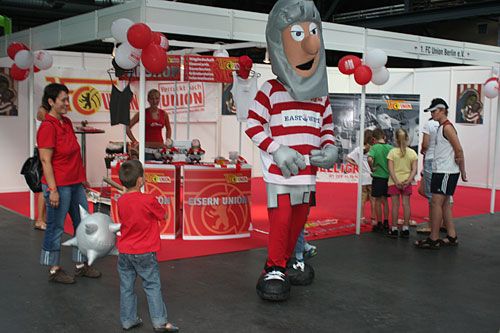 Ein großer Maskottchen-Kostümträger steht vor einem Stand mit roten und weißen Ballons, während Kinder und Erwachsene dort vorbeigehen.