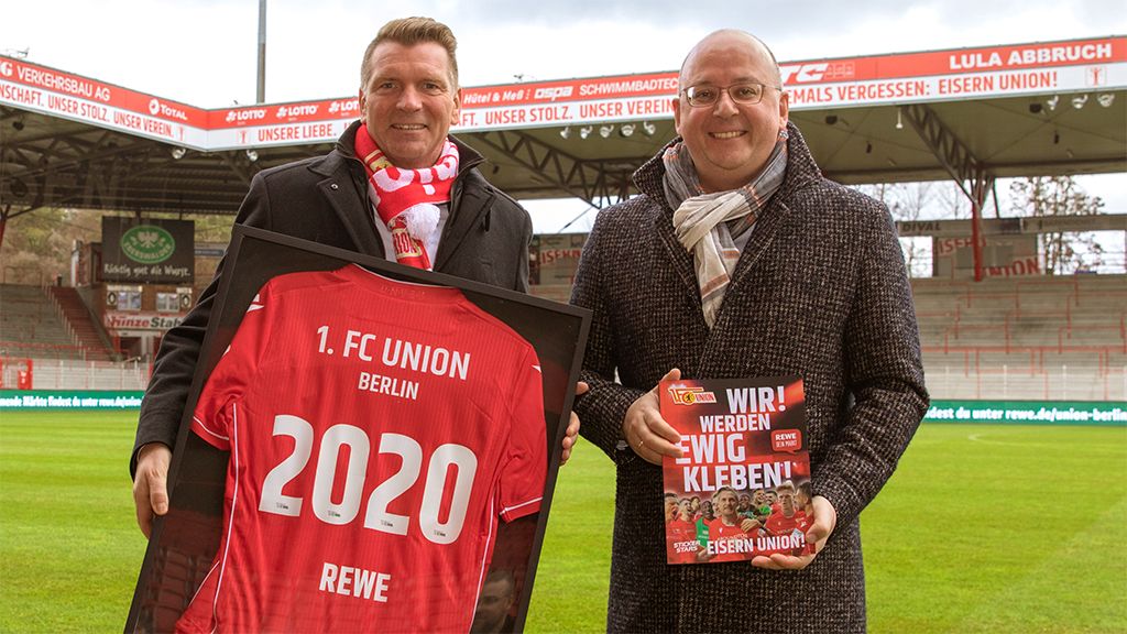 Two men are standing in the stadium of 1. FC Union Berlin, one holding a jersey and the other a magazine.