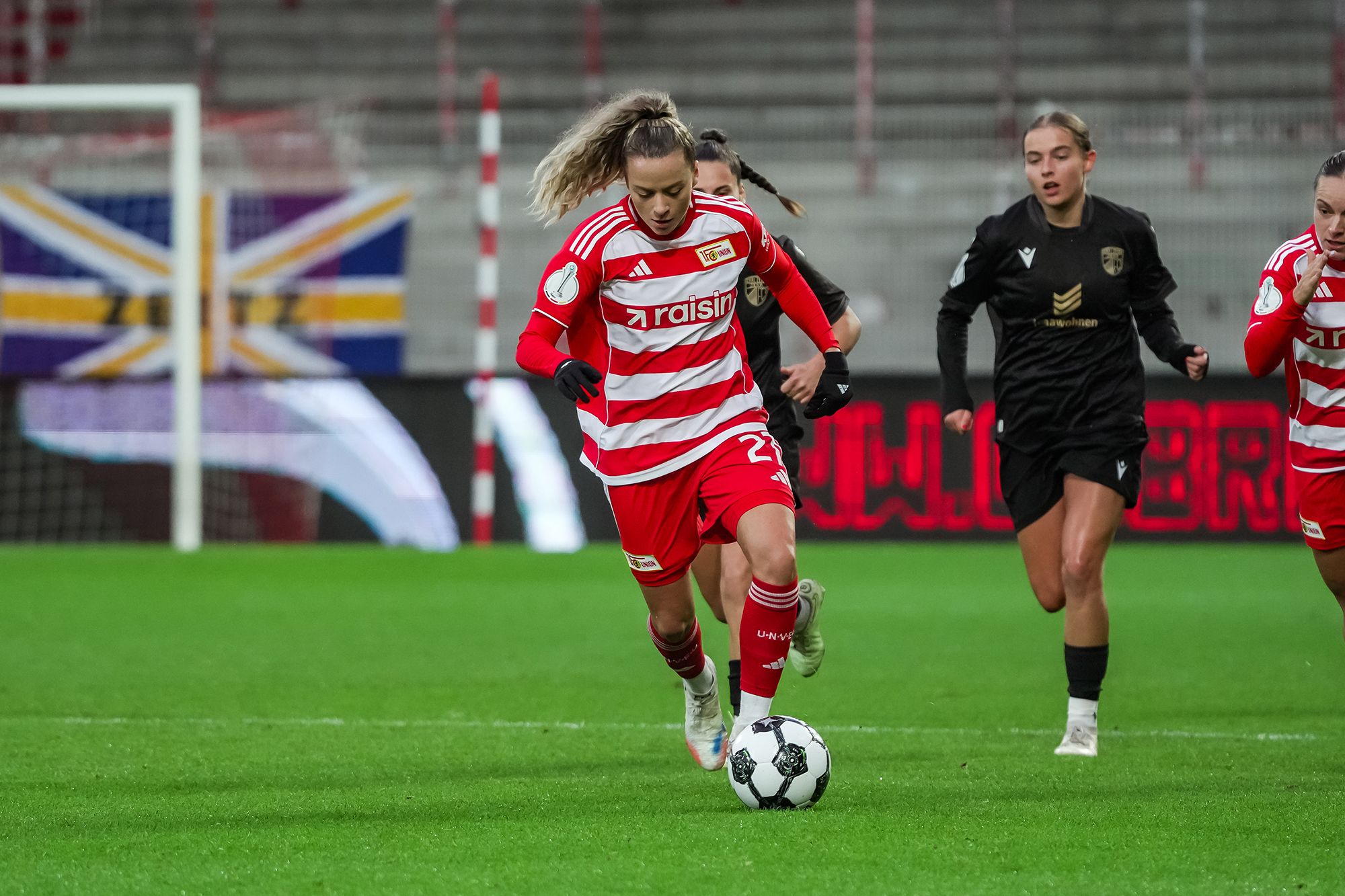 Eine Spielerin in roter und weißer Uniform rennt mit einem Fußball auf ein Tor zu, während andere Spielerinnen auf dem Spielfeld stehen.
