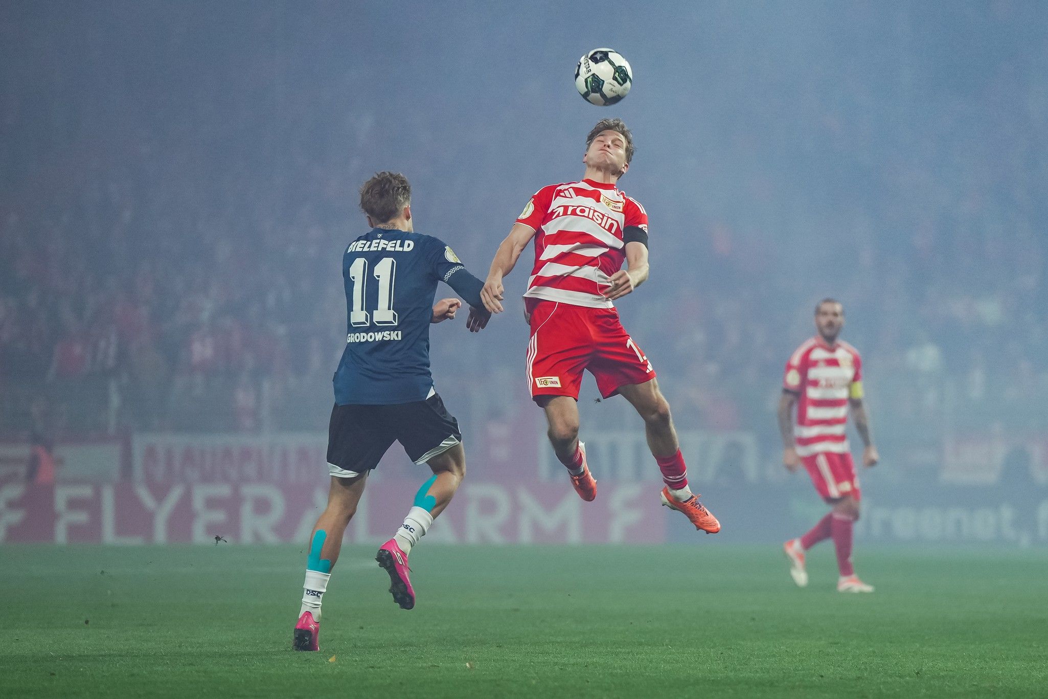 Spielszene im Fußball mit zwei Spielern in actionreichen Posen, während ein Ball in der Luft fliegt und Rauch im Hintergrund sichtbar ist.