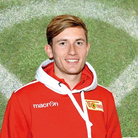 Lächelnder junger Mann in roter Sportjacke mit Kapuze vor einem Rasenplatz-Hintergrund.