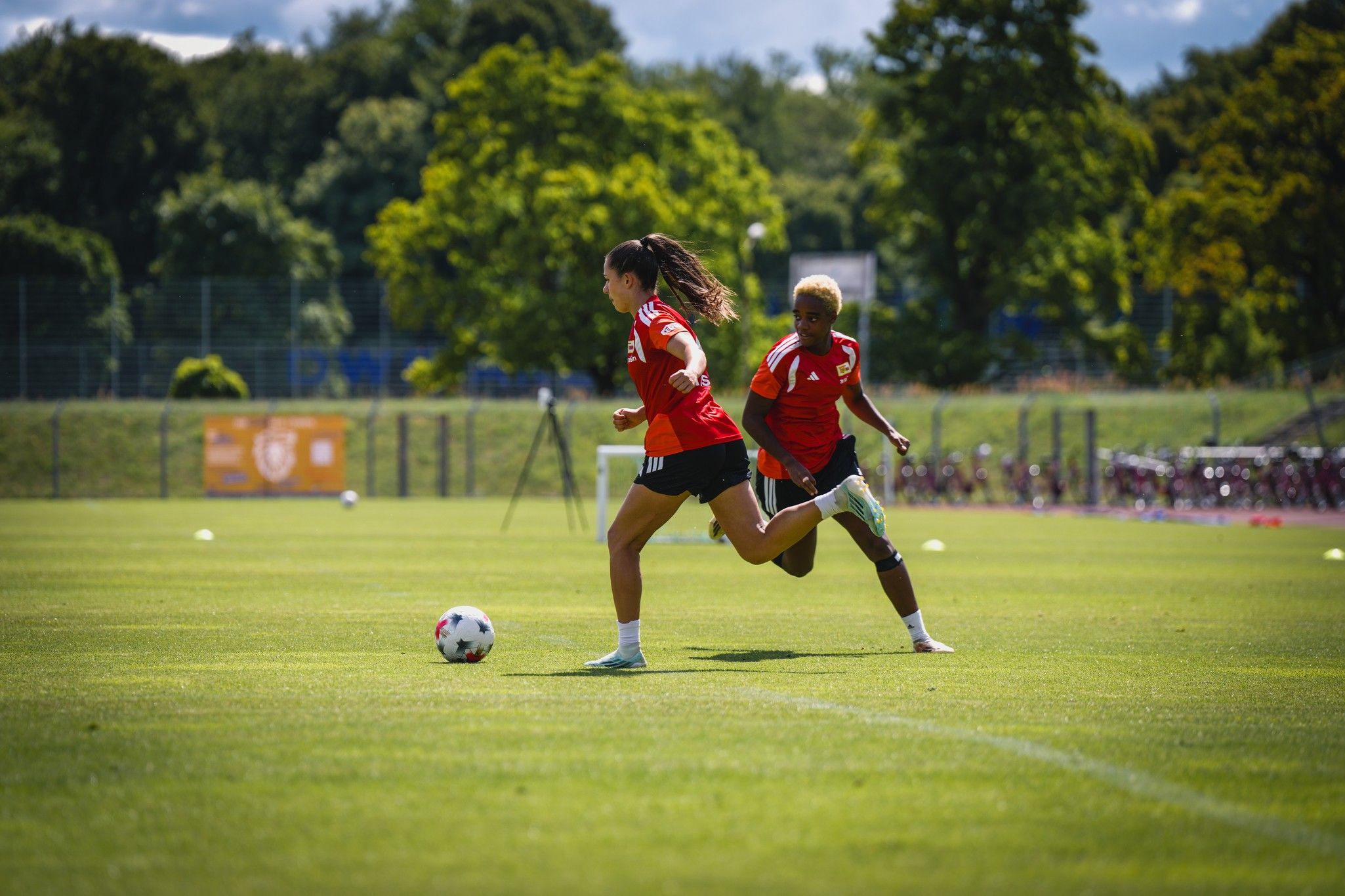 Zwei Fußballspielerinnen in roten Trikots sprinten über ein grünes Spielfeld, während ein Ball vor ihnen rollt.